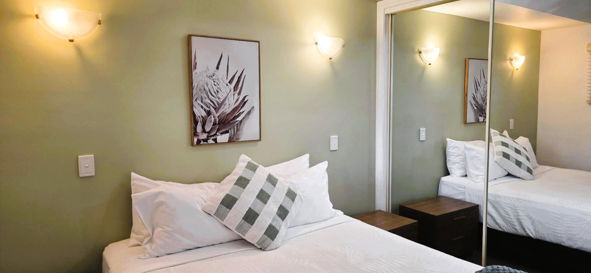 A hotel room with a bed, a bedside table, a mirror, and wall-mounted lights.