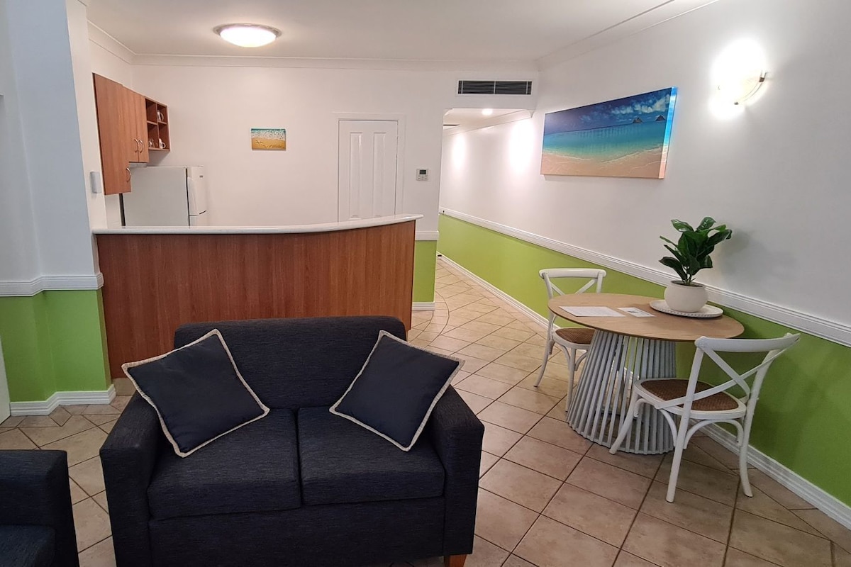 Una sala de estar con sofá y sillas, un área de cocina con armarios, una mesa de comedor con sillas y un cuadro en la pared.