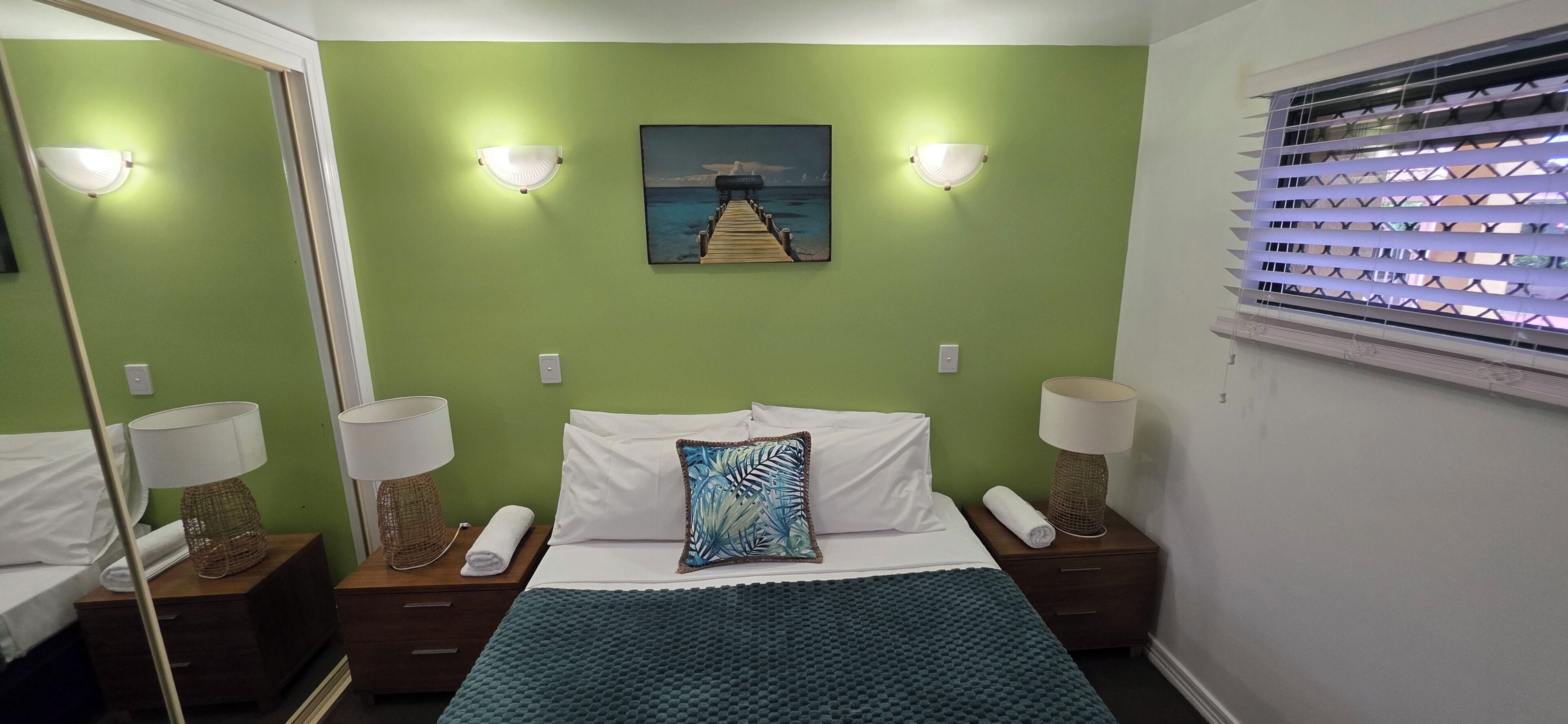 A hotel room with a bed, two bedside tables with lamps, a painting on the wall, and a window with blinds.