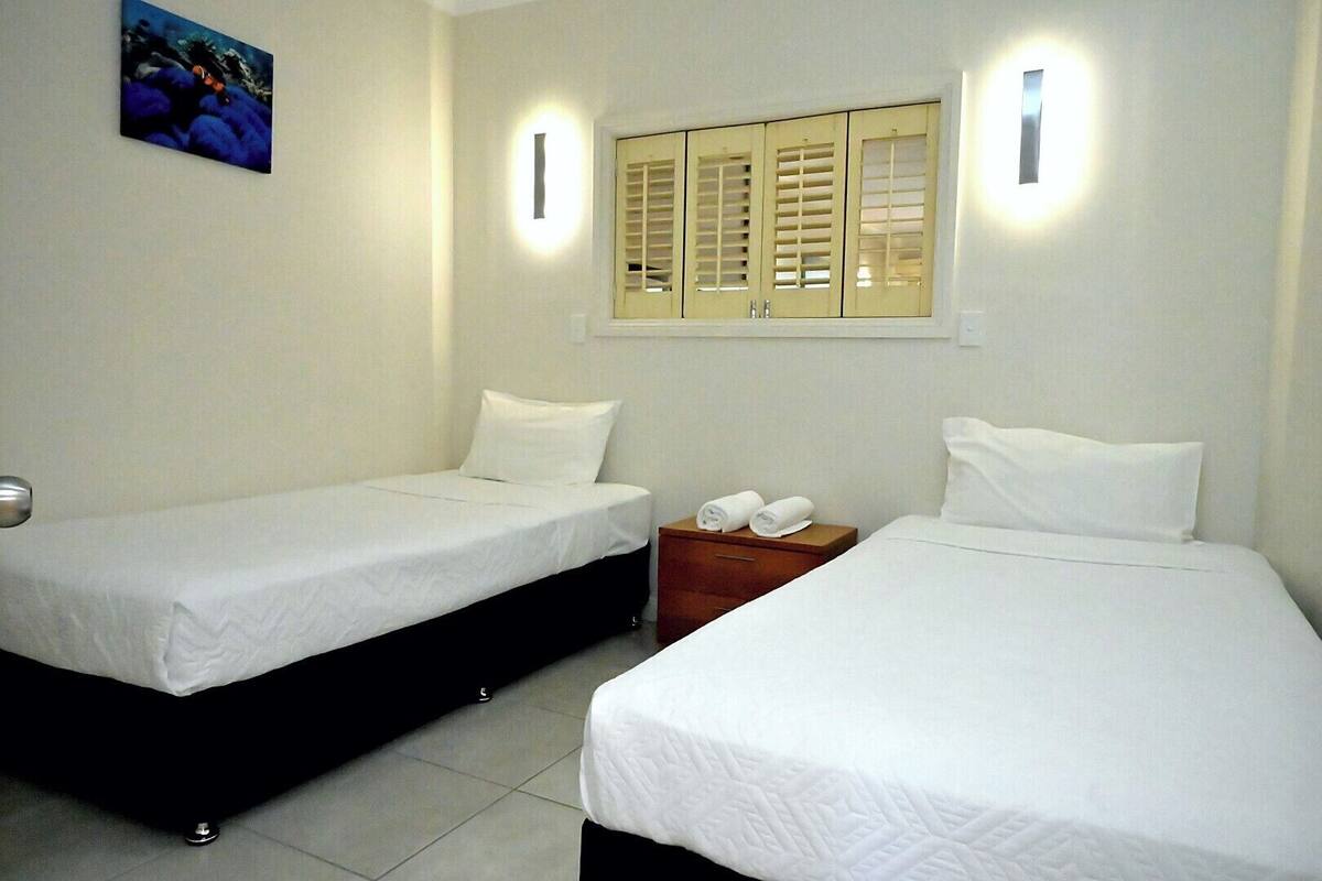 Two single beds with white linens, a nightstand with towels, a wall-mounted light, and a window with blinds.