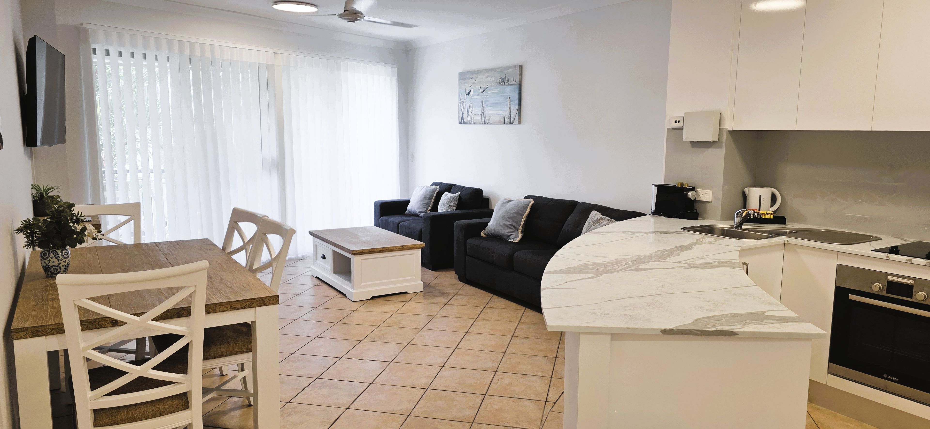 A modern kitchen with a built-in oven, a dining area with a wooden table and chairs, and a living area with a sofa and a painting on the wall.