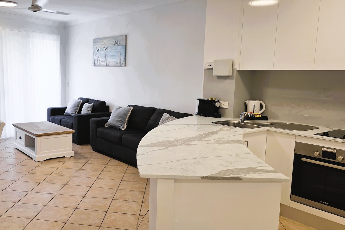 Una cocina moderna con horno empotrado, una zona de comedor con mesa y sillas de madera, y una sala de estar con sofá y un cuadro en la pared.