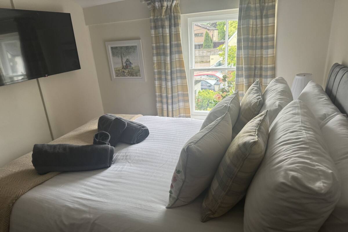 A neatly made bed with a white comforter and several pillows, a flat-screen TV mounted on the wall, a window with curtains, and a framed picture on the wall.