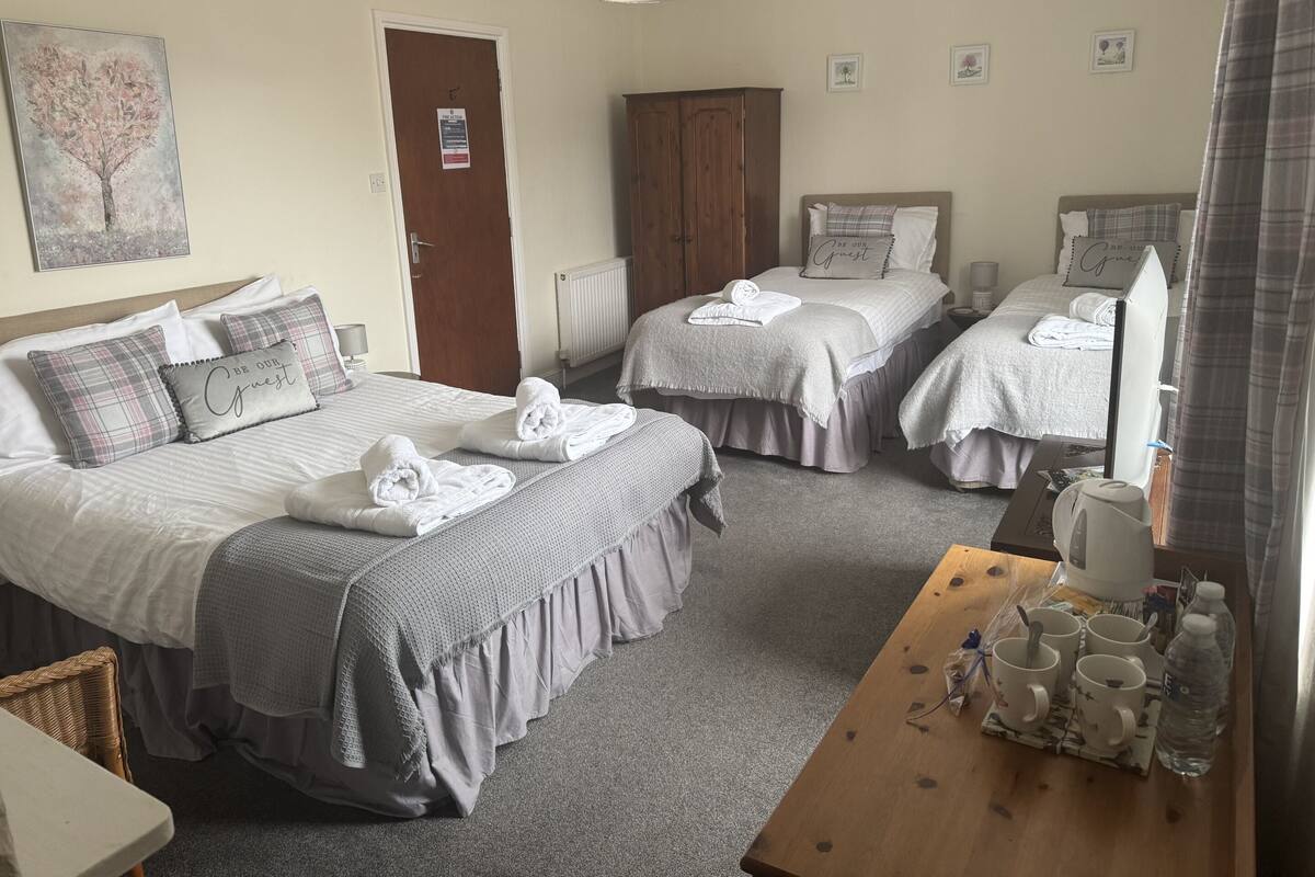 A hotel room with two beds, each with a pillow and a towel. There is a wooden wardrobe, a table with cups and a kettle, and a painting on the wall.