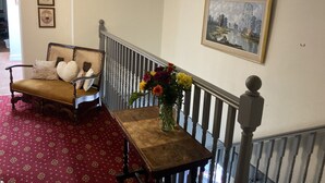 A room with a wooden bench, a small table with a vase of flowers, a framed painting on the wall, and a staircase with a railing. - Thorverton Arms (Exeter)