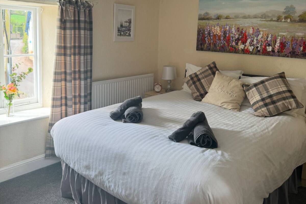 A neatly made bed with pillows and a blanket, a window with curtains, a radiator, a painting on the wall, and a lamp on the nightstand.