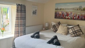 A neatly made bed with pillows and a blanket, a window with curtains, a radiator, a painting on the wall, and a lamp on the nightstand. - Thorverton Arms (Exeter)
