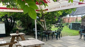 A patio area with wooden picnic tables and chairs, surrounded by greenery and a brick wall. - Thorverton Arms (Exeter)