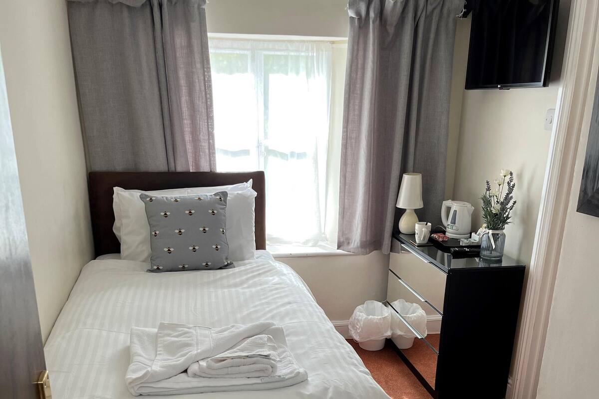 A neatly made bed with white linens, a bedside table with a lamp, a small cabinet, and a window with curtains.