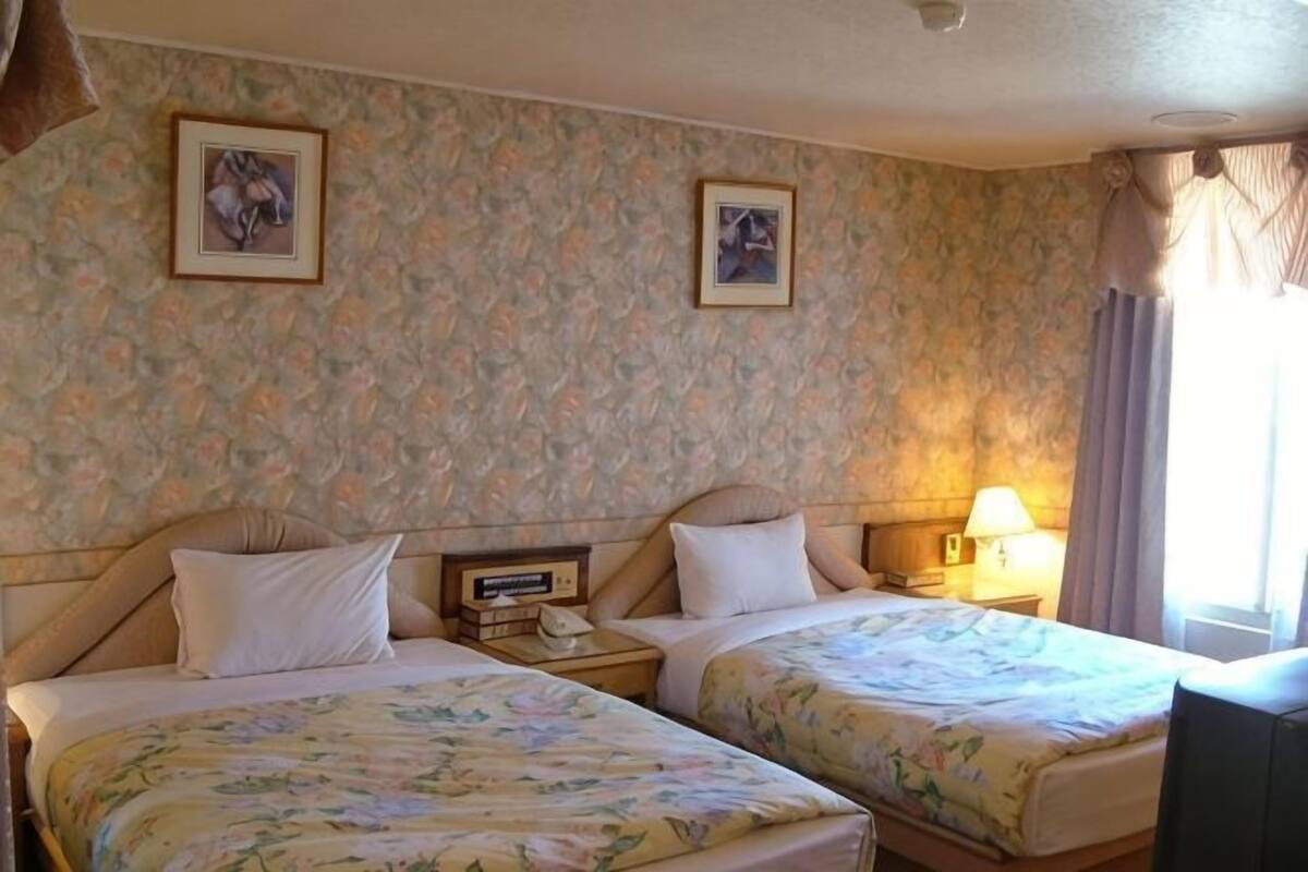 Een hotelkamer met twee bedden, bloemenbehang, ingelijste foto’s aan de muur, bedlampjes en een raam met gordijnen.