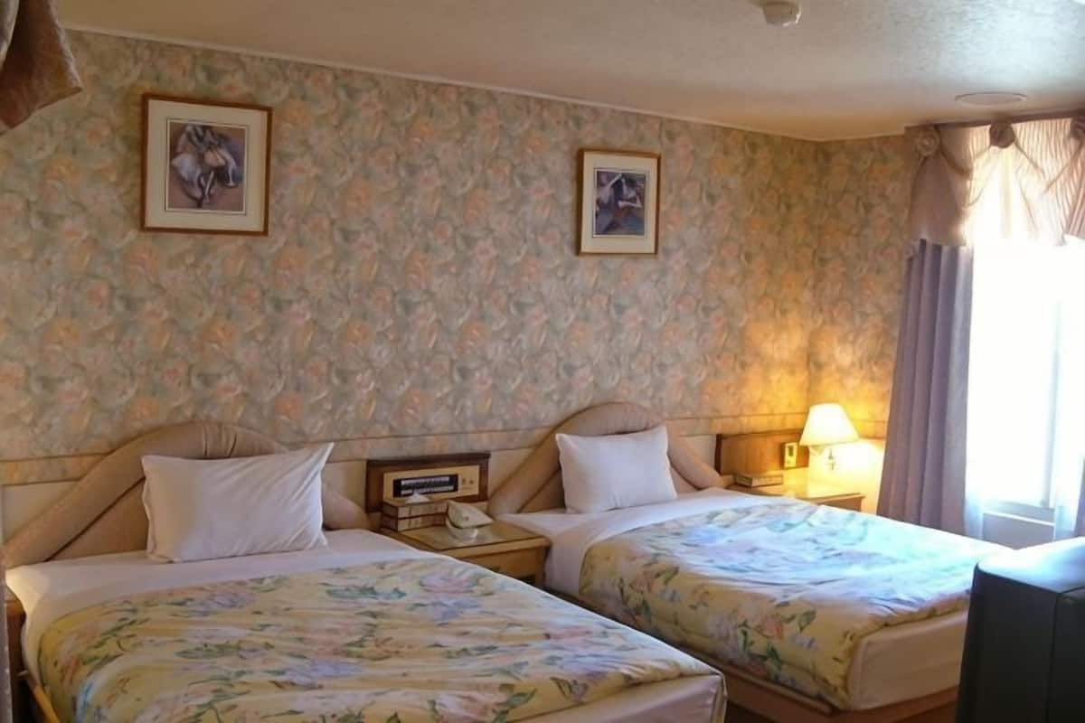 Ein Hotelzimmer mit zwei Betten, floralem Wandpapier, gerahmten Bildern an der Wand, Nachttischlampen und einem Fenster mit Vorhängen.