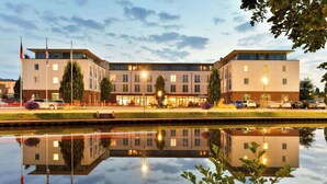 A modern hotel with a reflection in a calm body of water, featuring a symmetrical facade, multiple windows, and a flat roof. - Best Western Plus Hotel Papenburg (Papenburg)