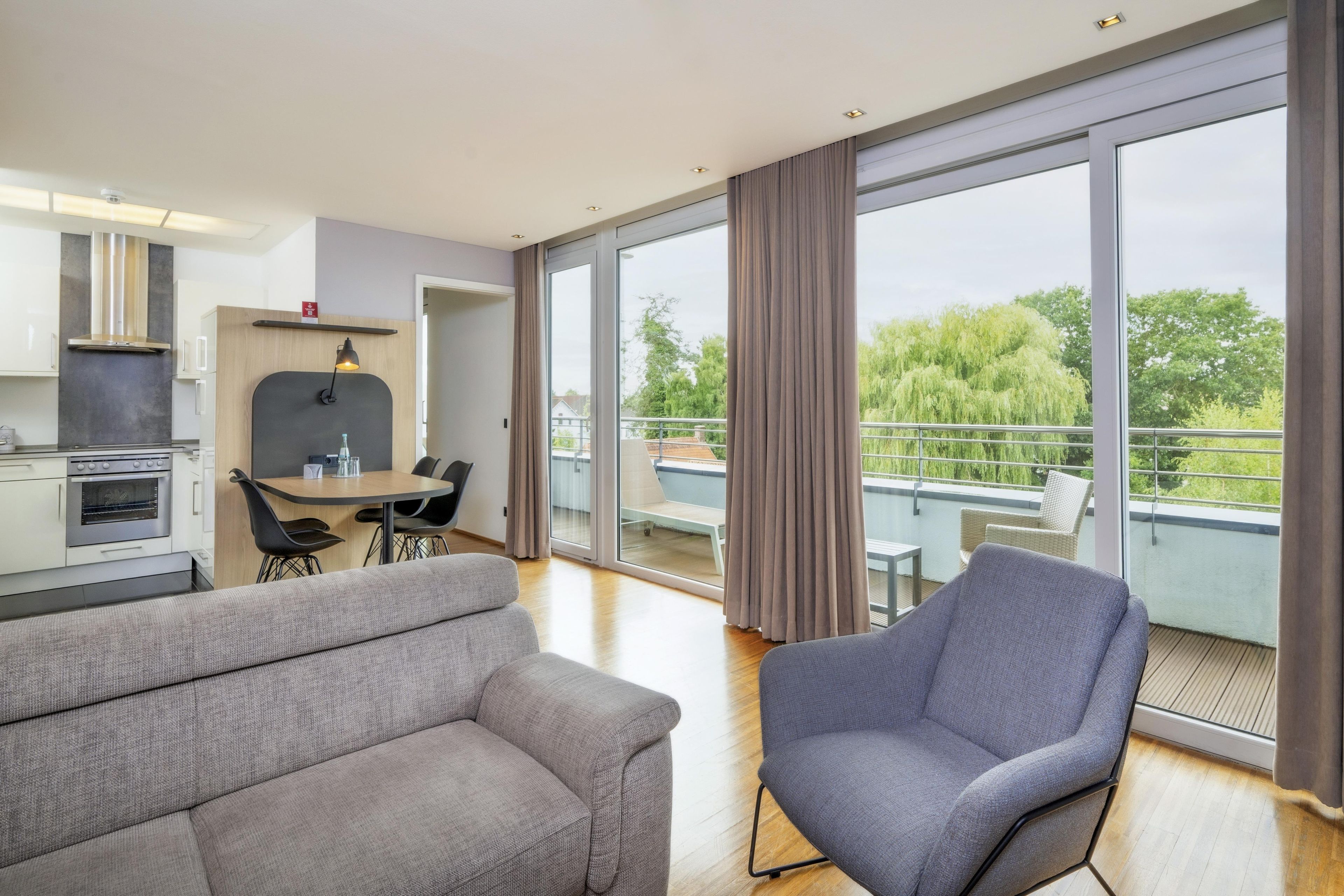 Una sala de estar moderna con sofá, una mesa de comedor y una silla. Hay una zona de cocina con estufa y alacenas. La habitación tiene ventanas grandes con cortinas y vistas a áreas verdes.