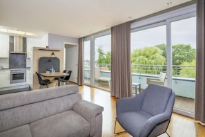 A modern living room with a sofa, a dining table, and a chair. There is a kitchen area with a stove and cabinets. The room has large windows with curtains and a view of greenery outside. - Best Western Plus Hotel Papenburg (Papenburg)