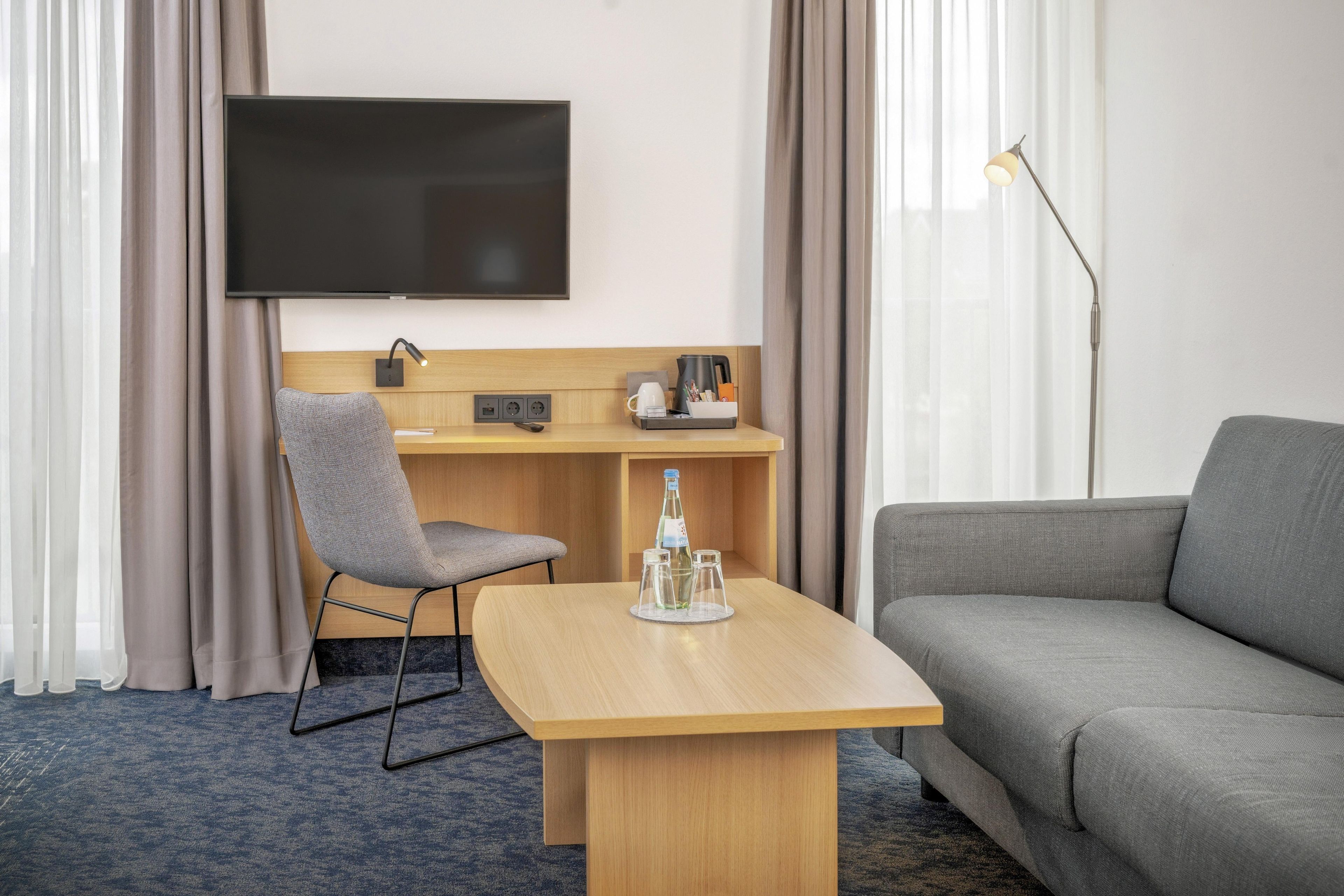 Hotellihuone, jossa on seinälle asennettu taulutelevisio, työpöytä tuolilla, sohva ja lamppu.