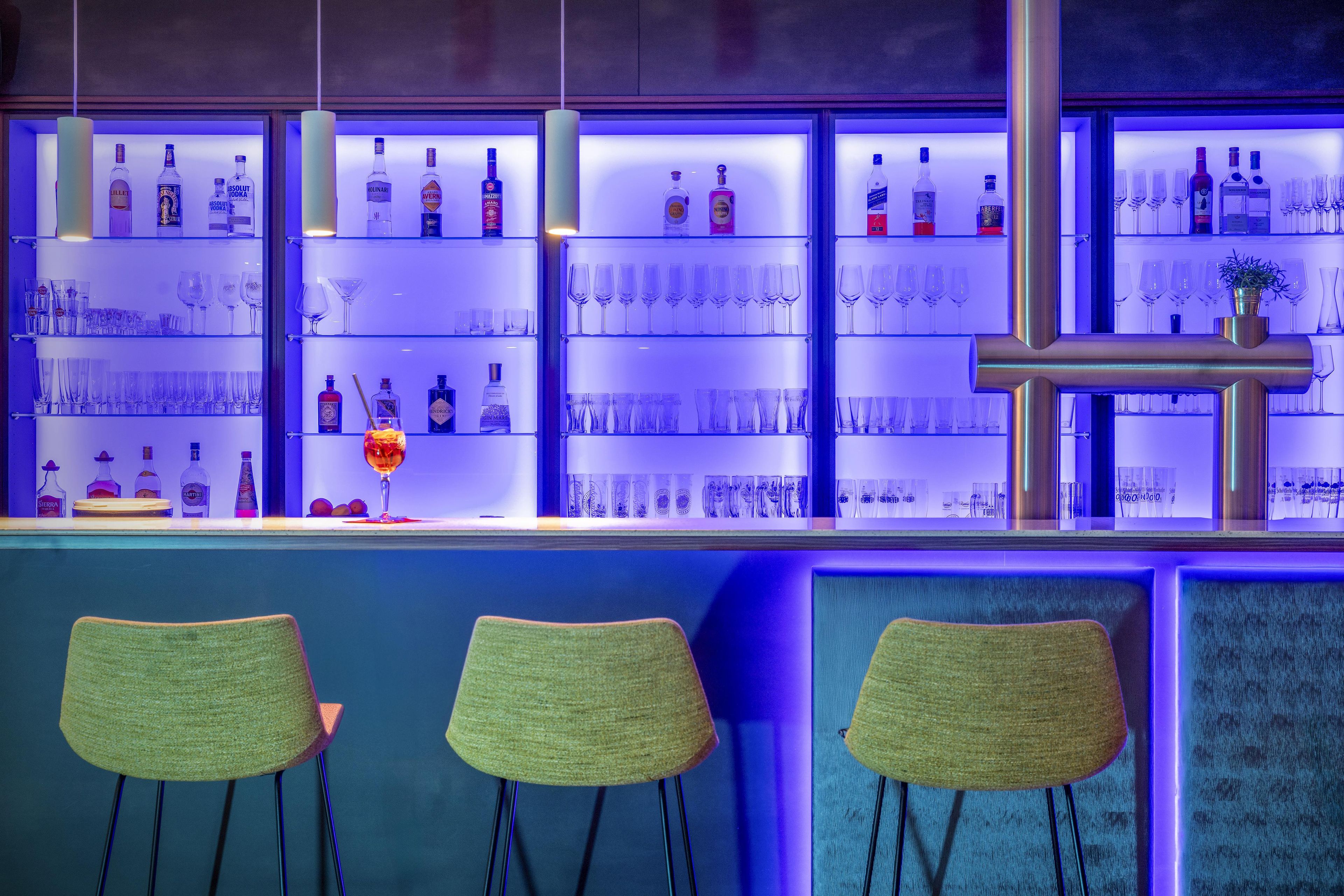 Eine Bar mit mehreren Regalen voller Spirituosenflaschen und Gläsern sowie drei grünen Barhocker.