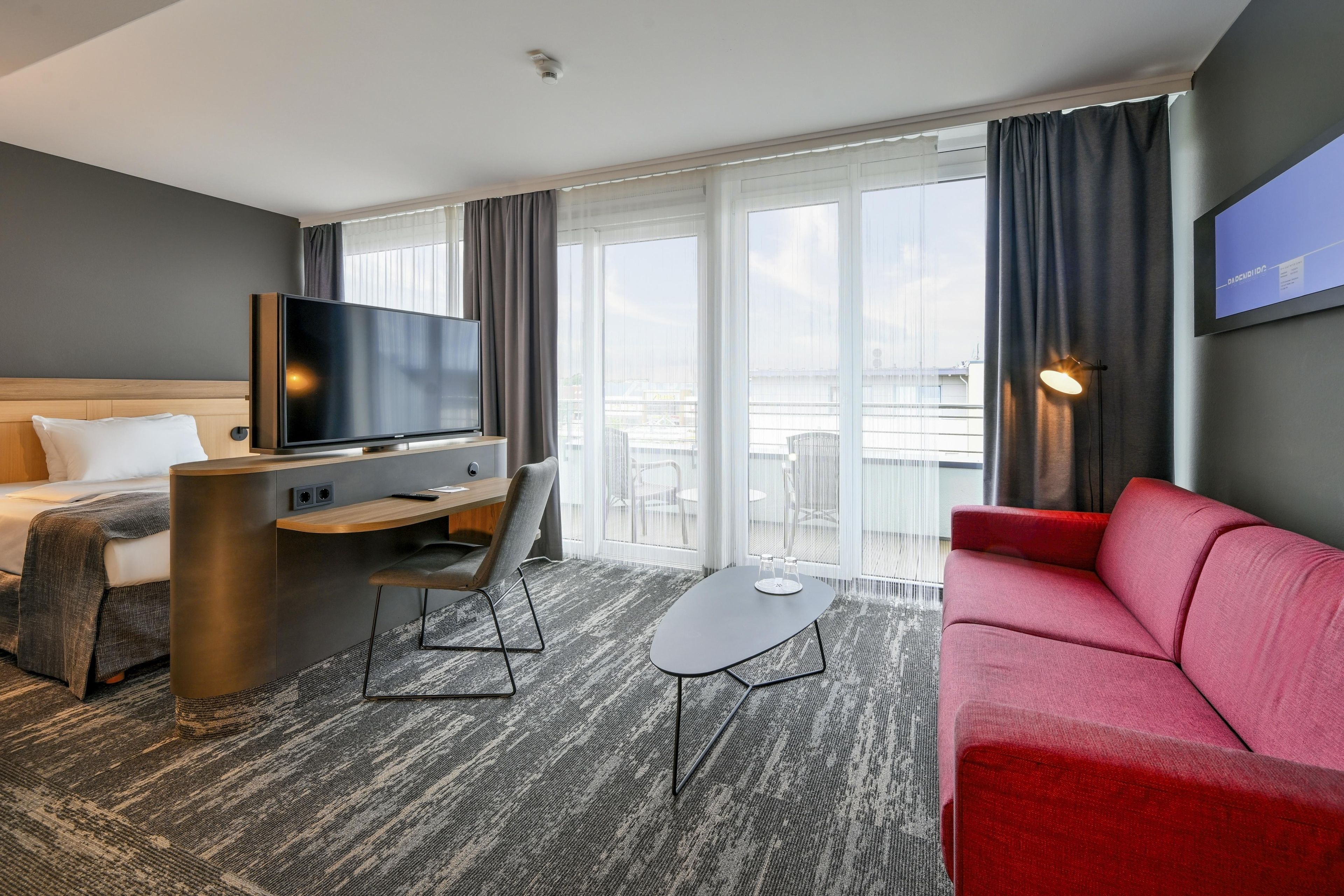 Ein Hotelzimmer mit Bett, Schreibtisch, Fernseher, einem roten Sofa und einem Balkon mit Blick.