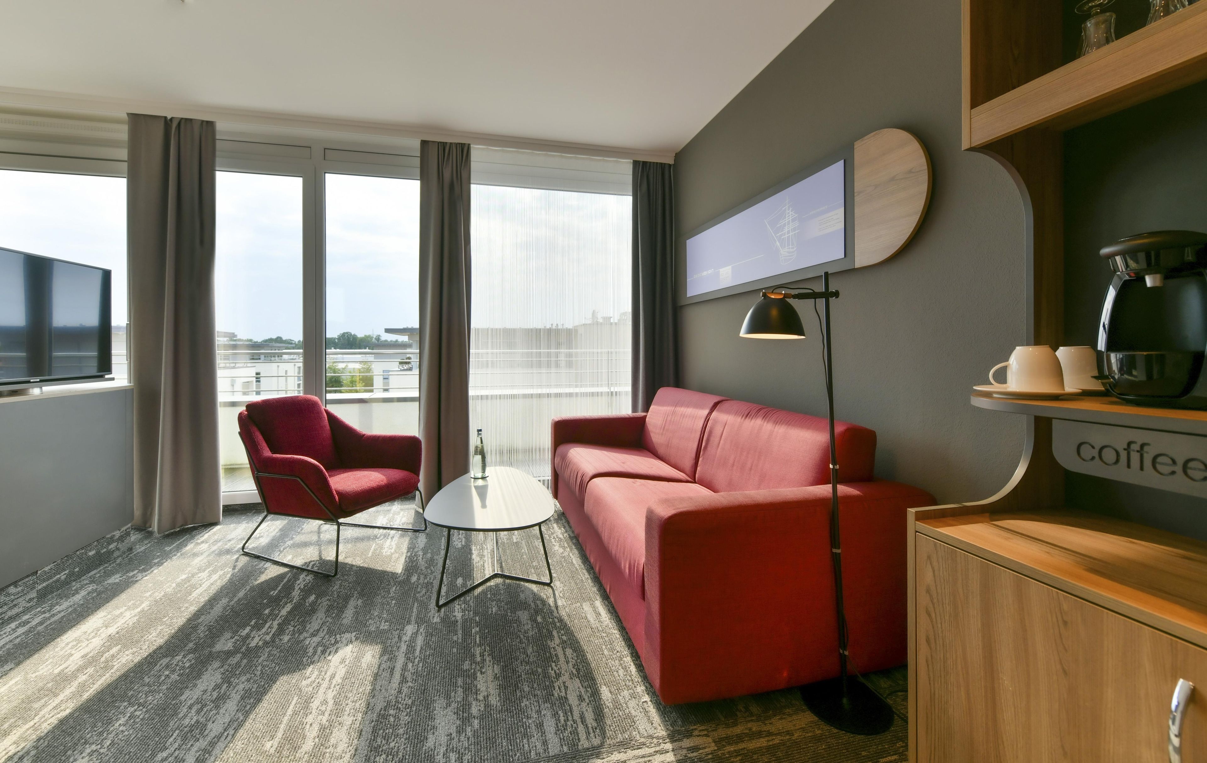 Ein modernes Hotelzimmer mit einem roten Sofa, einem roten Sessel, einem kleinen runden Tisch, einer Kaffeemaschine und einem an der Wand befestigten Fernseher.