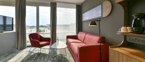 Habitación de hotel moderna con un sofá rojo, un sillón rojo, una mesita redonda pequeña, una cafetera y un televisor montado en la pared.