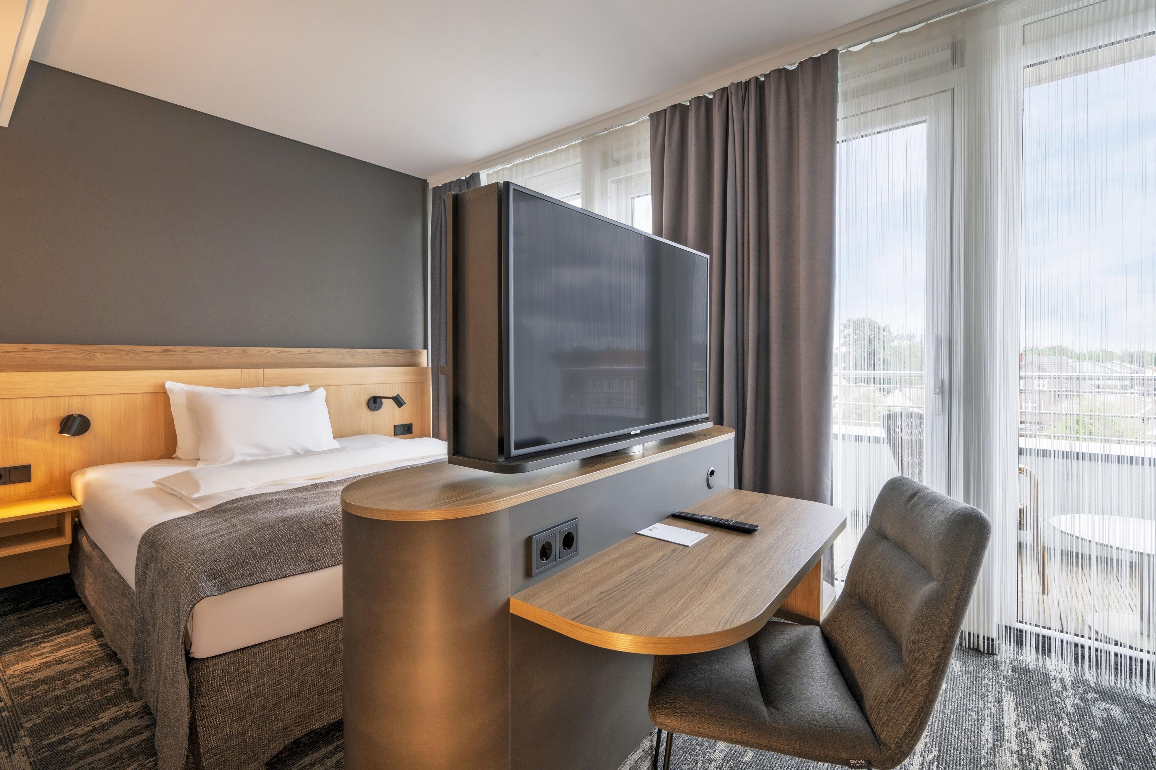 Ein Hotelzimmer mit einem Bett, einem Fernseher auf einem Schrank, einem Schreibtisch mit Stuhl und Blick auf die Stadt durch große Fenster.
