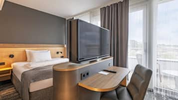 Ein Hotelzimmer mit einem Bett, einem Fernseher auf einem Schrank, einem Schreibtisch mit Stuhl und Blick auf die Stadt durch große Fenster.