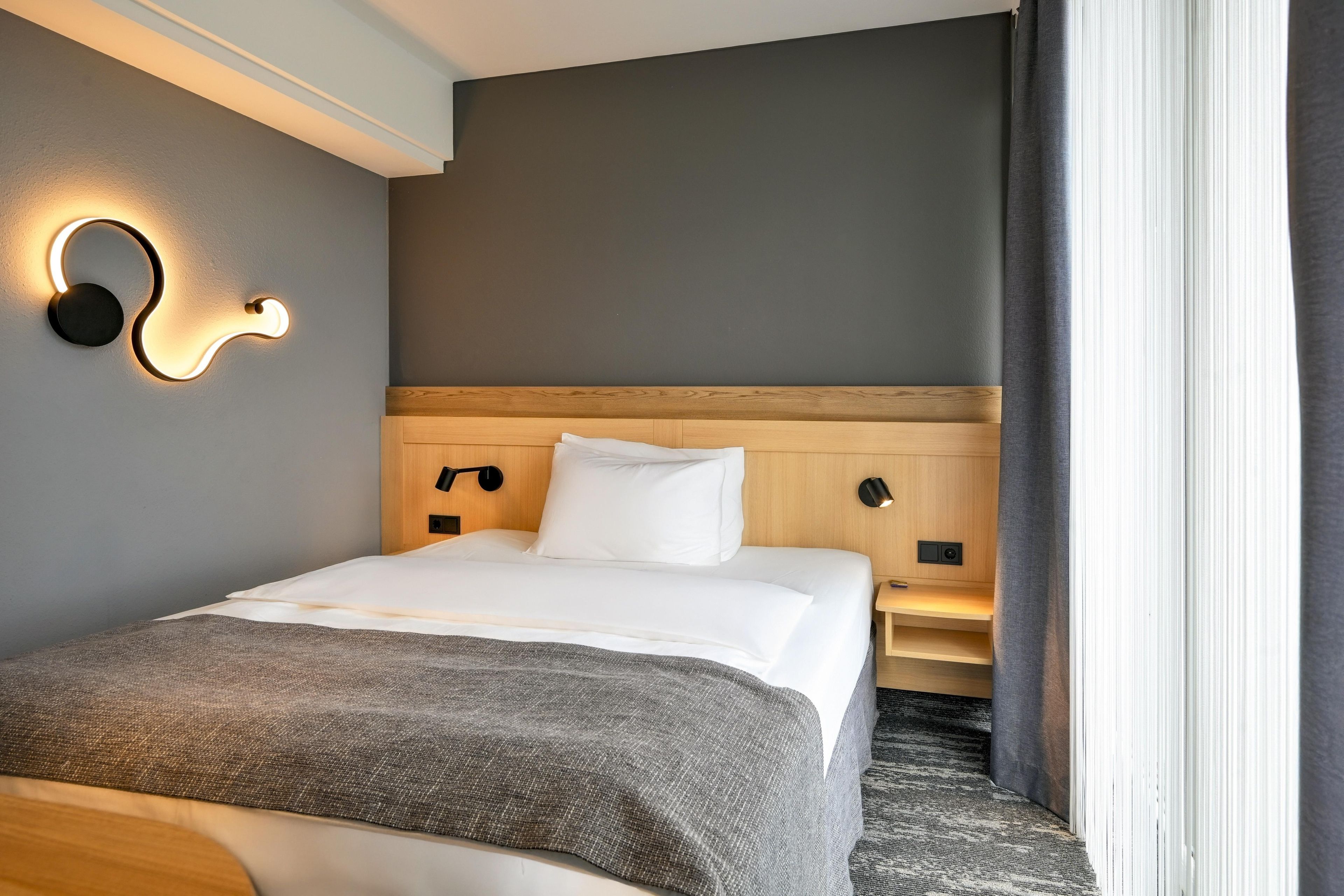 Una habitación de hotel con cama, lámparas de noche, cabecera y ventana con cortinas.