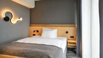 Una habitación de hotel con cama, lámparas de noche, cabecera y ventana con cortinas.