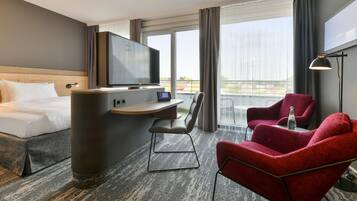 Une chambre d’hôtel avec un lit, un bureau, un téléviseur à écran plat, une chaise, un fauteuil rouge et une grande fenêtre.