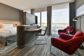 Ein Hotelzimmer mit einem Bett, einem Schreibtisch, einem Flachbildfernseher, einem Sessel sowie einem roten Ohrensessel und einem großen Fenster.