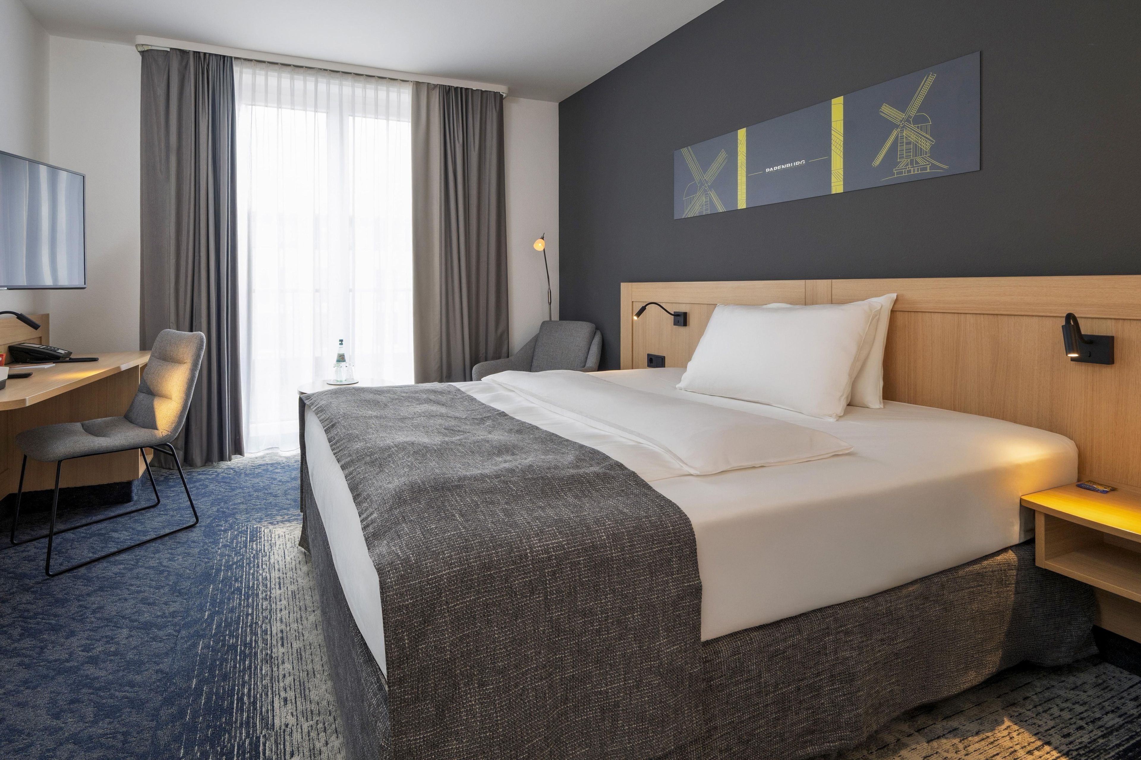 A hotel room with a large bed, a desk with a chair, a TV, and a window with curtains.