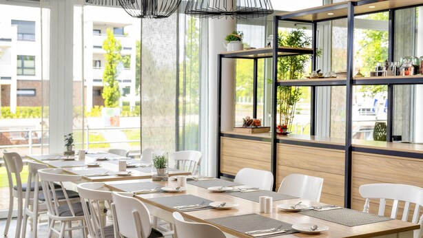A modern dining area with a long table set for six, white chairs, and a glass partition with shelves. - Best Western Plus Hotel Papenburg (Papenburg)