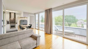 A modern living room with a sofa, dining table, and chairs, a kitchen with a stove and cabinets, and large sliding glass doors to a balcony with a view of the neighborhood. - Best Western Plus Hotel Papenburg (Papenburg)