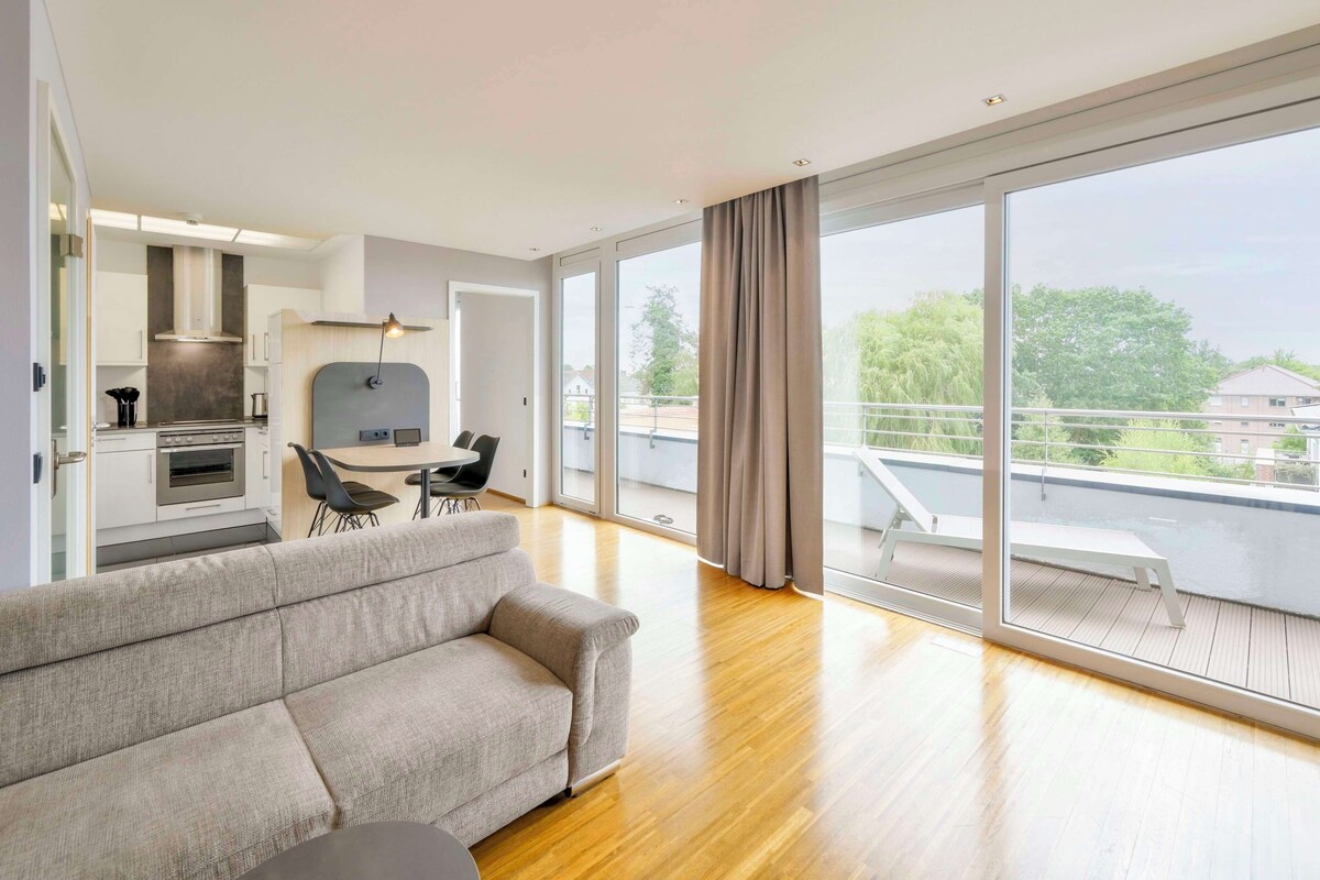 Een moderne woonkamer met een bank, eettafel en stoelen, een keuken met fornuis en kastjes, en grote schuifpuien naar een balkon met uitzicht op de buurt.