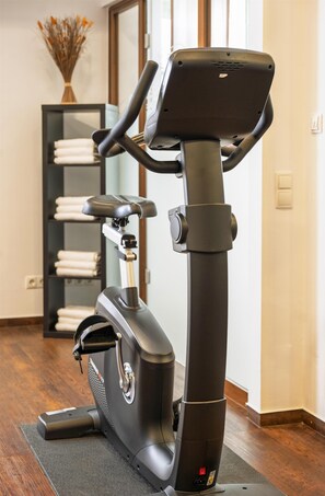 Una bicicleta estática en una habitación con piso de madera, un estante con toallas dobladas y un jarrón con flores secas.