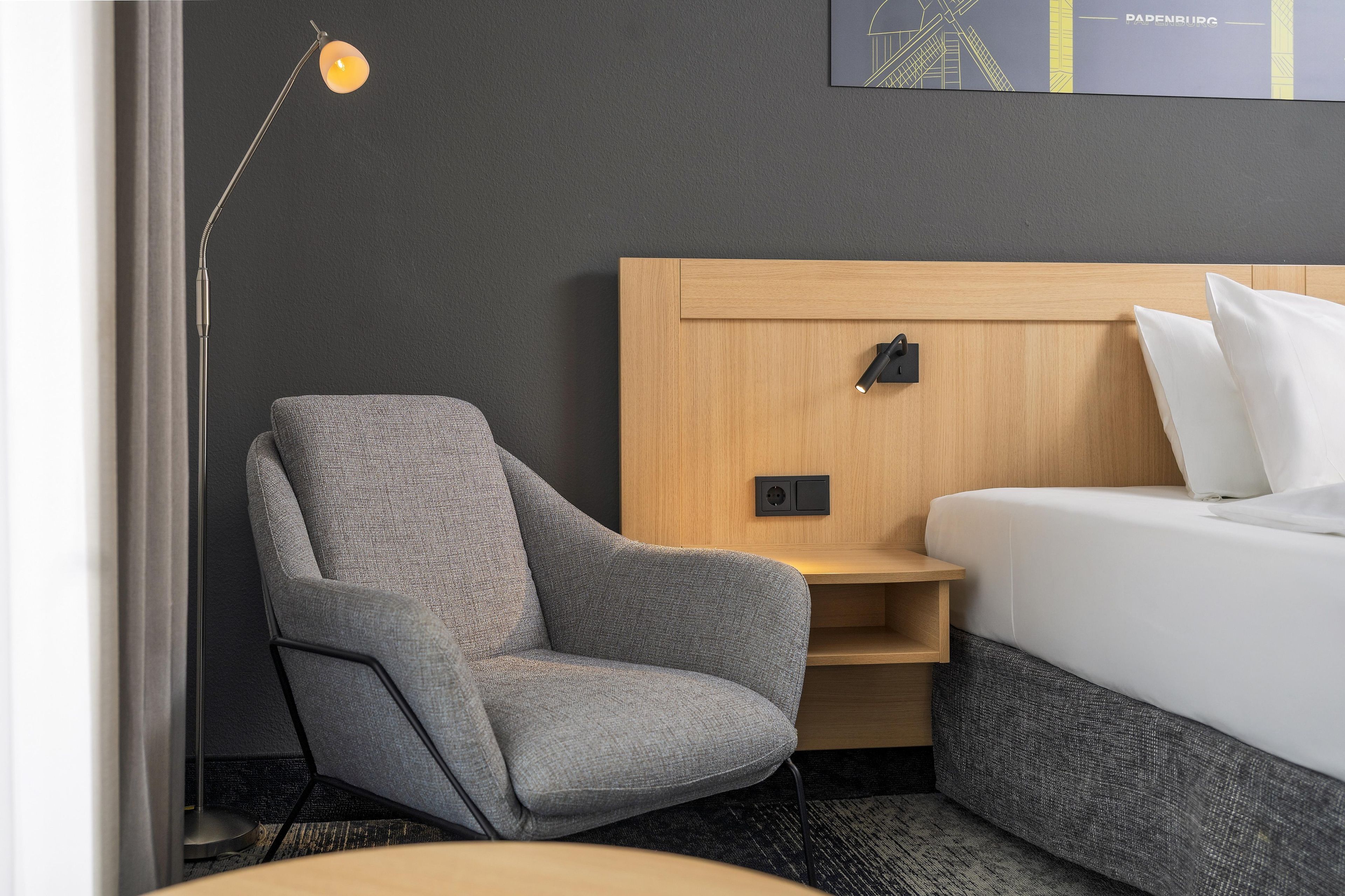 Une chambre d’hôtel avec un fauteuil gris, une tête de lit en bois, un lit blanc et une lampe sur pied.