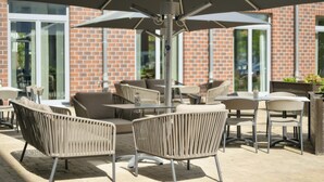 An outdoor patio with wicker furniture, a central umbrella, and a brick building in the background.