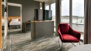 A modern hotel room with a bed, a TV on a stand, a red armchair, and large windows offering a city view. - Best Western Plus Hotel Papenburg (Papenburg)