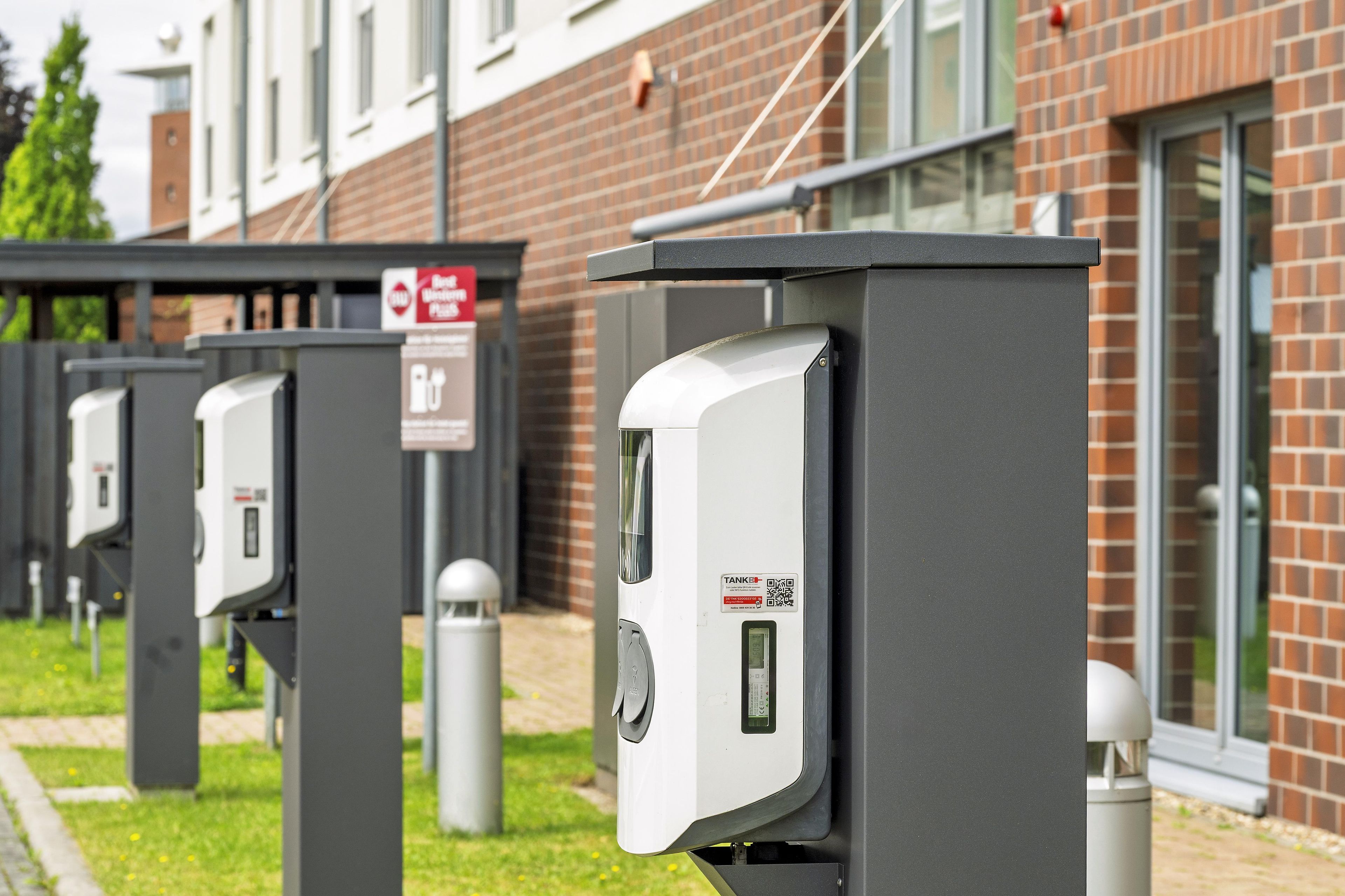 Eine Reihe von Ladestationen für Elektrofahrzeuge im Freien, mit einem Backsteingebäude im Hintergrund.