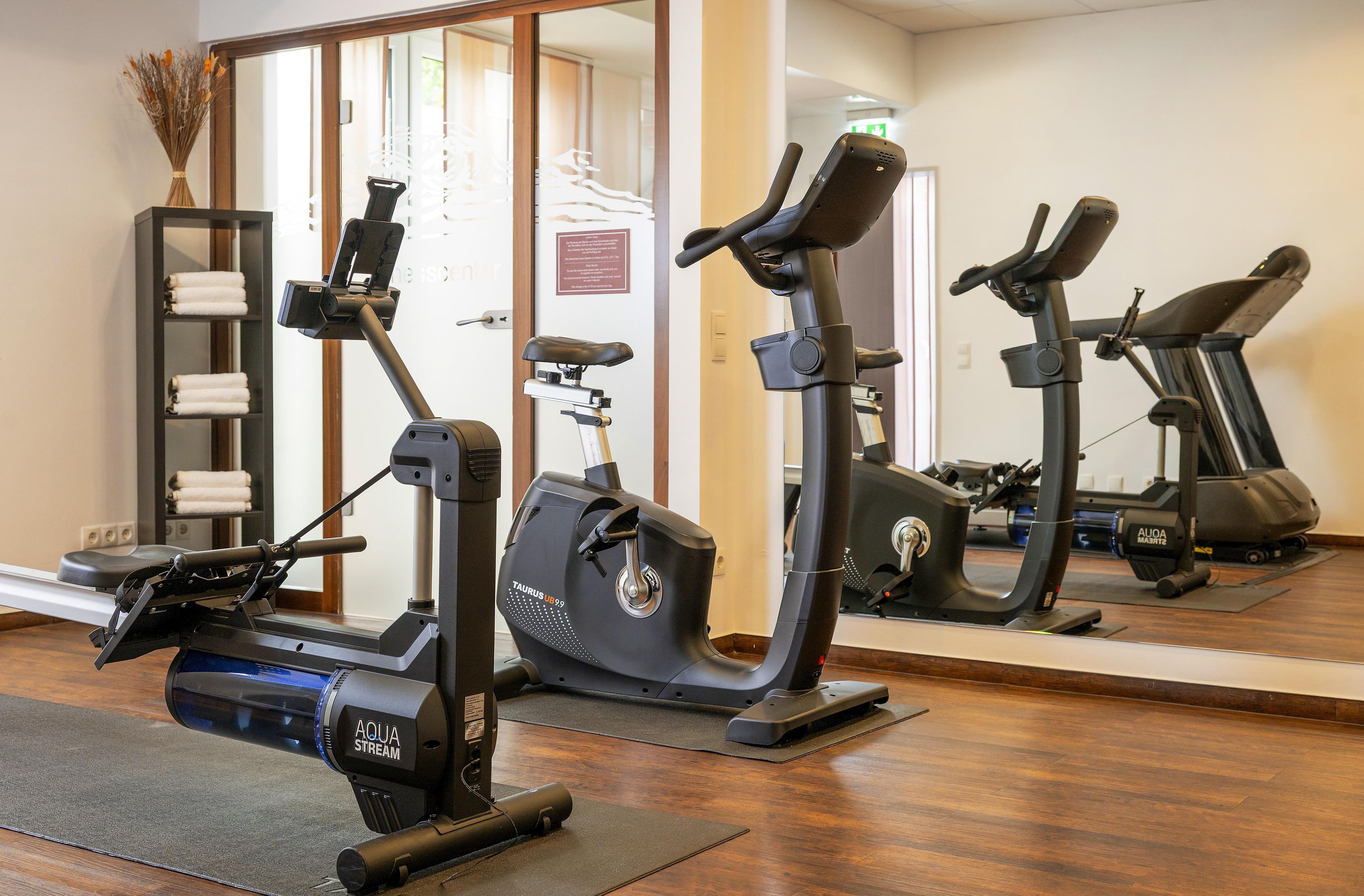 Un gimnasio con bicicletas estáticas y una cinta de correr, suelo de madera y un espejo que refleja la sala.