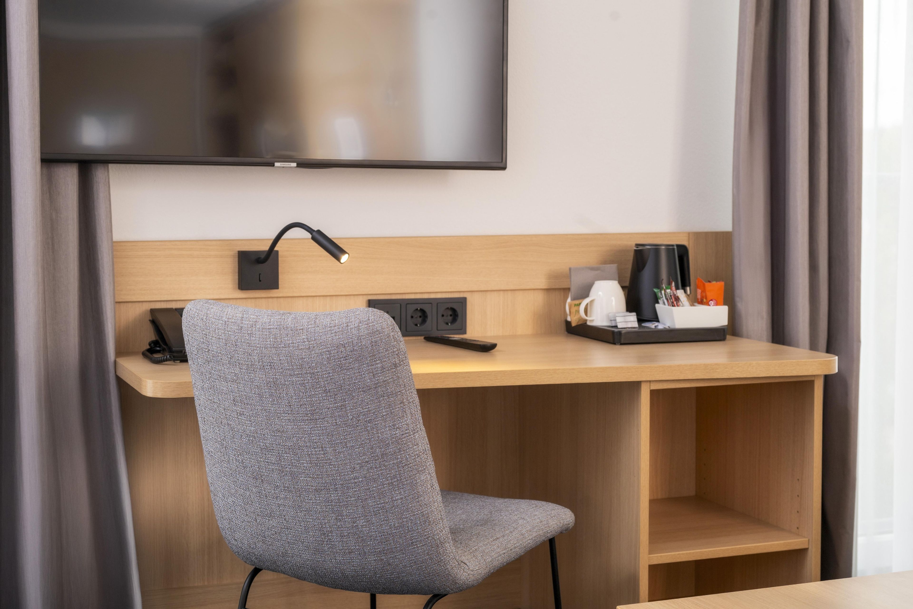 Habitación de hotel con un escritorio de madera, una butaca tapizada en gris, un televisor de pantalla plana montado en la pared, un enchufe y una cafetera sobre el escritorio.