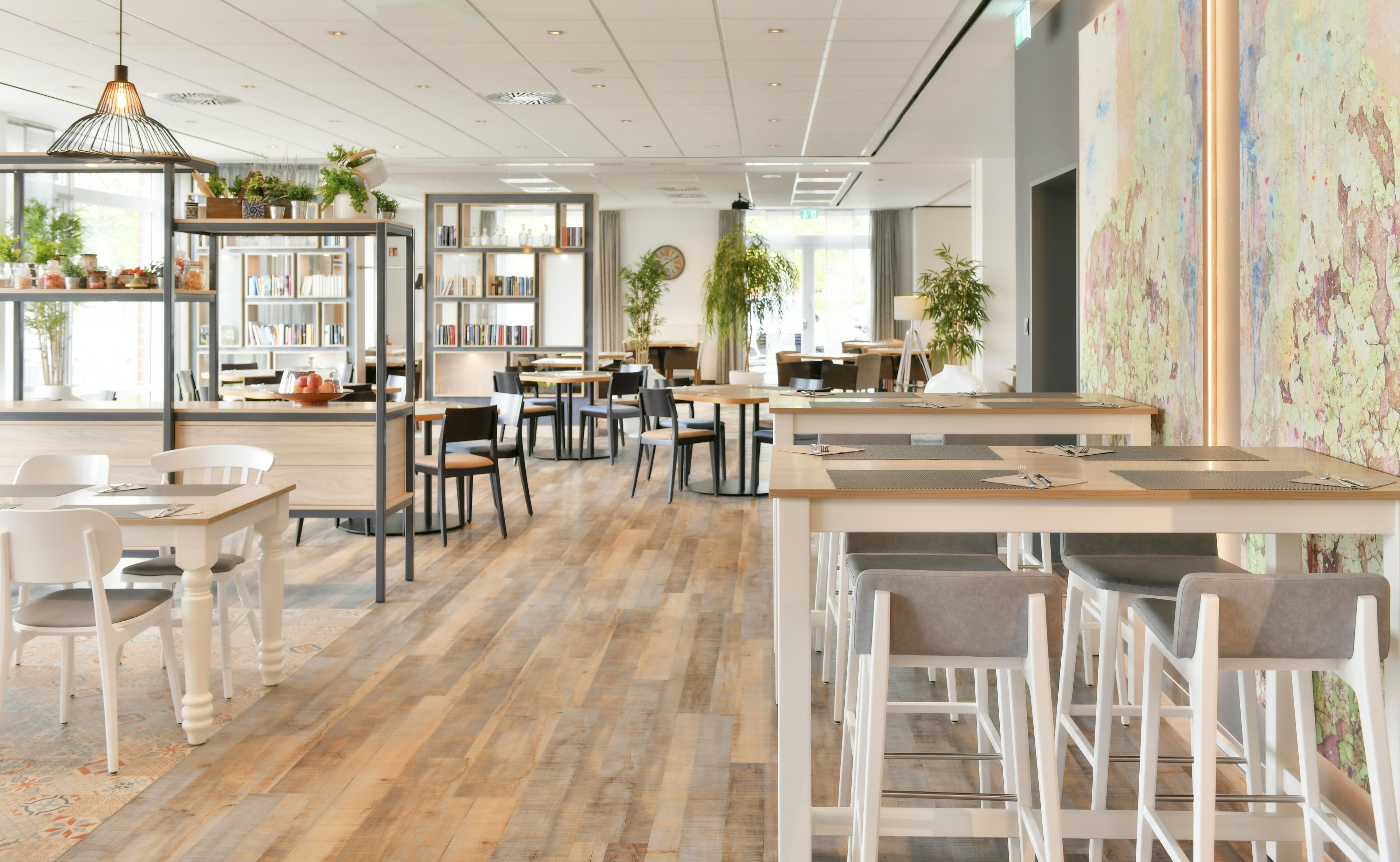 Un café moderno con suelo de madera, sillas y mesas blancas. Se observan estanterías y plantas al fondo.