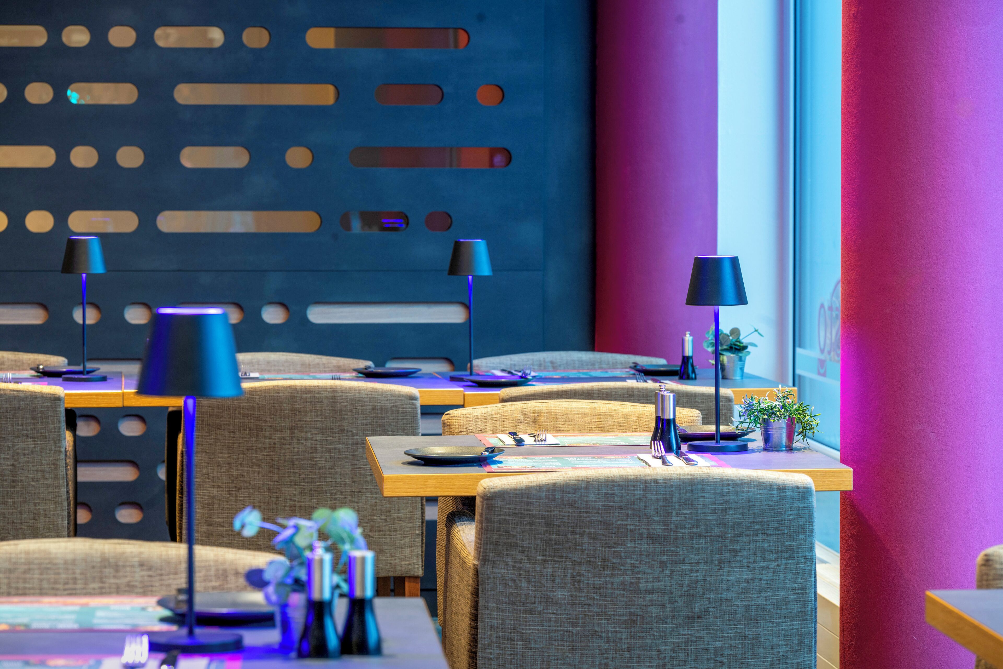 A modern restaurant with multiple tables and chairs, blue and purple lighting, and a decorative wall with cut-out patterns.