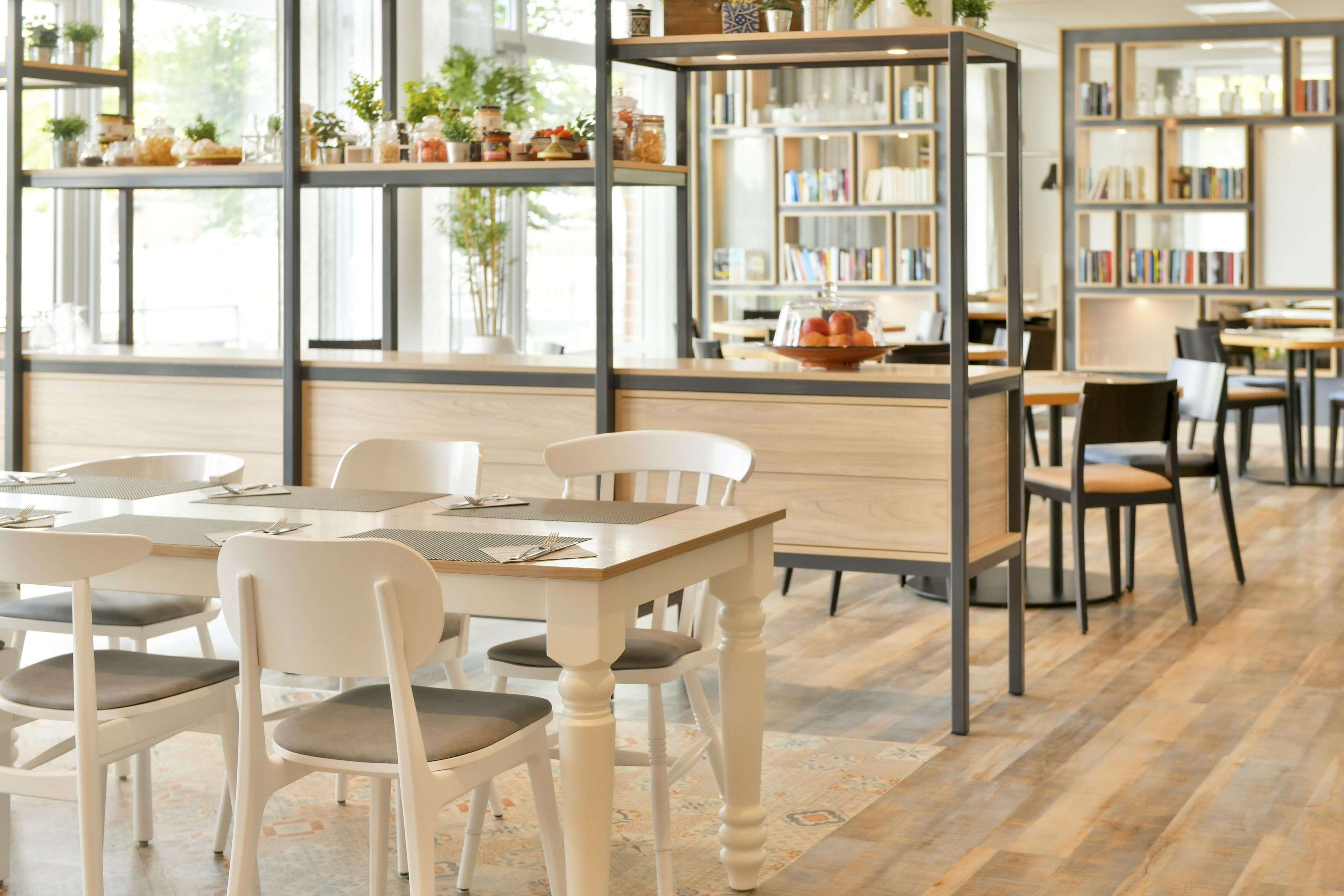 Interior de una cafetería con mesas, sillas, una estantería y un mostrador con vitrina.