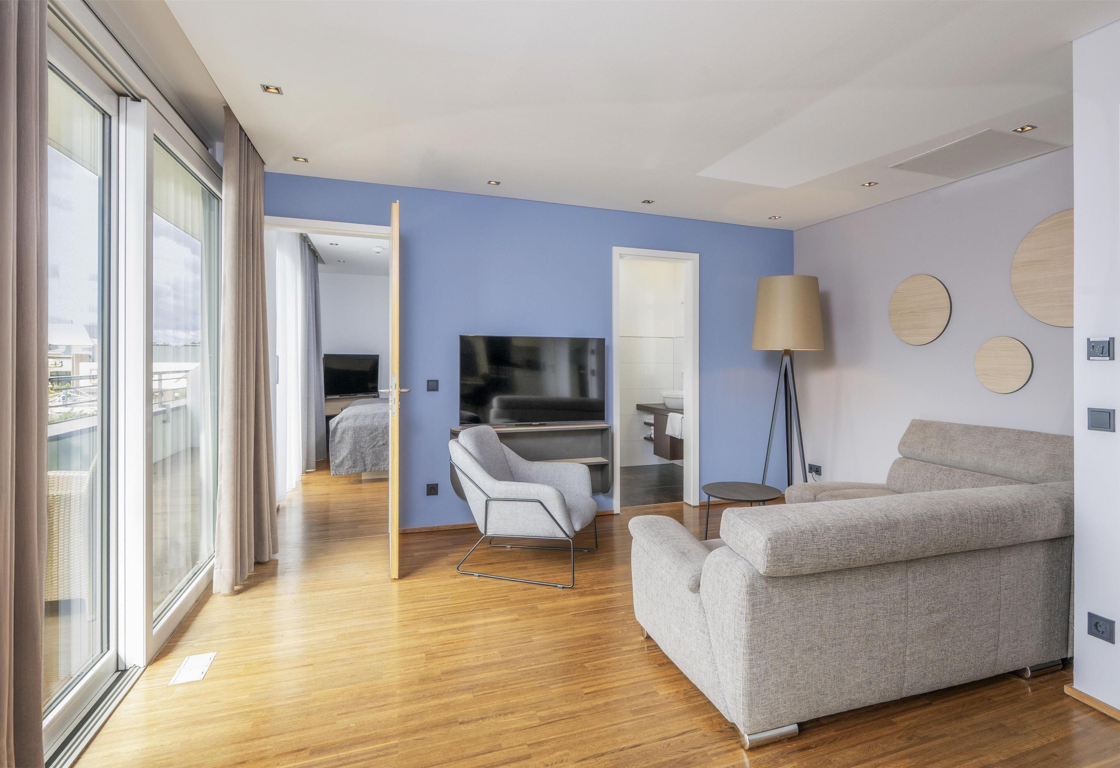 A modern hotel room with a wooden floor, a grey sofa, a TV, and a large window with curtains.