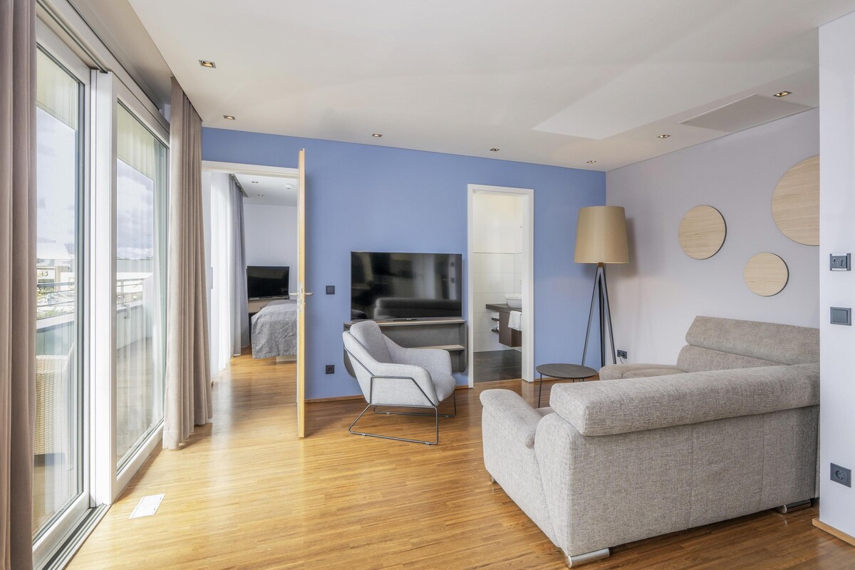 A modern hotel room with a wooden floor, a grey sofa, a TV, and a large window with curtains.