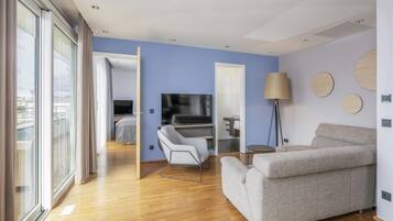 A modern hotel room with a wooden floor, a grey sofa, a TV, and a large window with curtains.