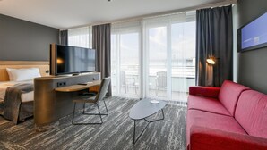 A hotel room with a bed, a TV, a desk, a red sofa, and large windows offering a view of the city. - Best Western Plus Hotel Papenburg (Papenburg)