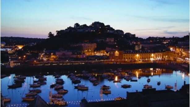 Une ville côtière au crépuscule, avec des bateaux amarrés dans un port, des bâtiments illuminés et une construction située au sommet d’une colline.
