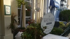 Une enseigne indiquant « The Wayfarer Guest House », avec une télévision dans toutes les chambres, un tableau encadré au mur et des plantes en pot disposées autour de l’entrée.