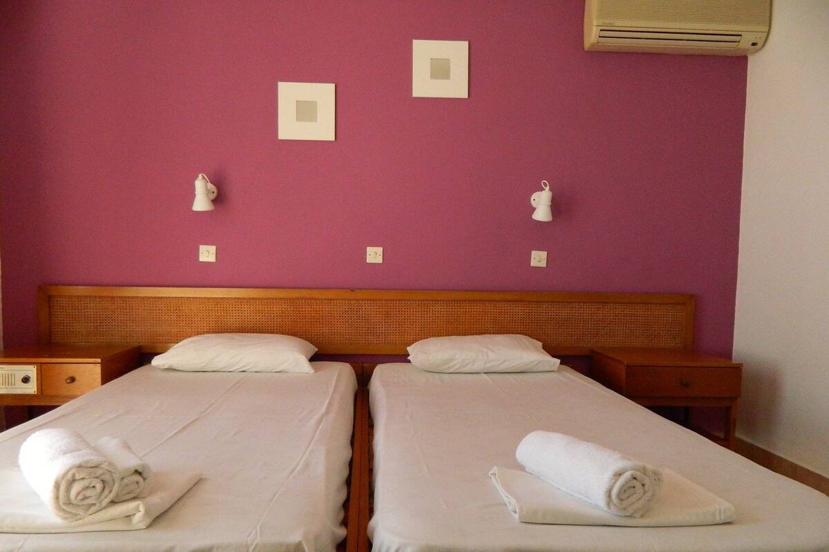Une chambre double avec deux lits simples, des têtes de lit en bois, du linge de lit blanc et un mur violet.
